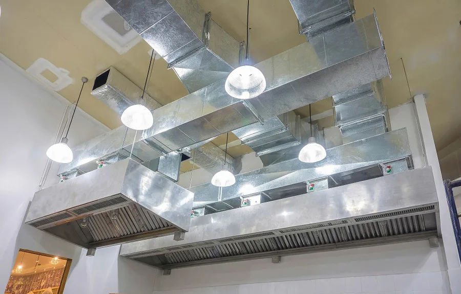 Stainless steel commercial kitchen hood with exposed exhaust ductwork running across the ceiling