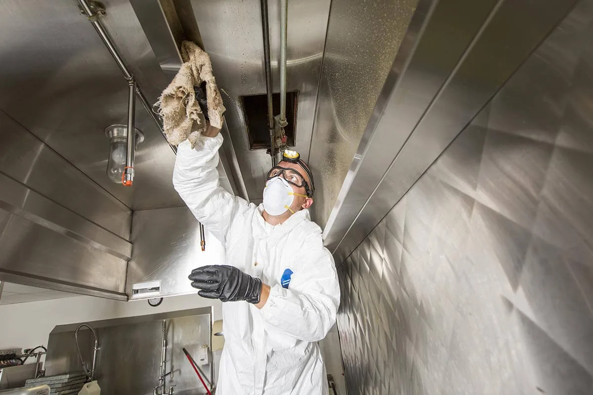 Technician in protective coveralls scraping grease from the inside of a commercial kitchen hood plenum